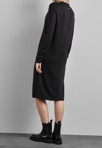 Robe noire à manches longues avec une coupe décontractée, dotée d'un détail de bouton à l'arrière, associée à des bottines noires. Texture de tissu lisse.