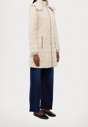 Femme portant une longue veste matelassée beige avec poches zippées, un jean large foncé, et des ballerines noires, debout devant un fond blanc.