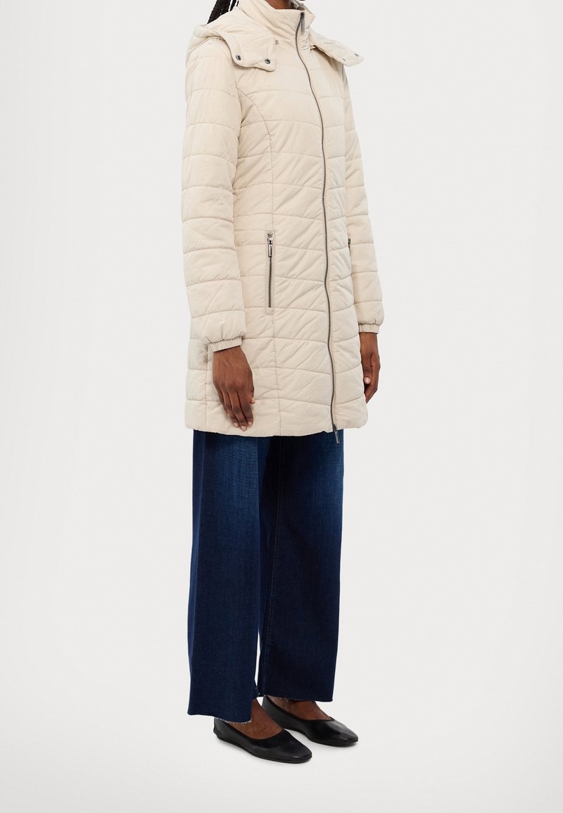 Femme portant une longue veste matelassée beige avec poches zippées, un jean large foncé, et des ballerines noires, debout devant un fond blanc.