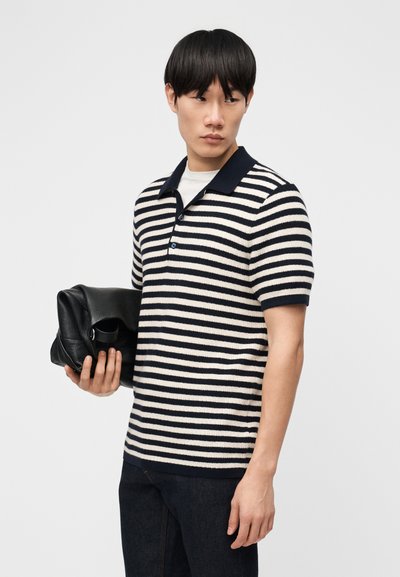 Mladý muž v černobílém polo tričku s vodorovnými pruhy, držící složenou černou koženou tašku, dívající se stranou.