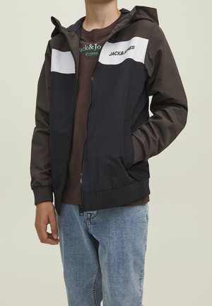 Persona che indossa una giacca Jack & Jones nera e grigia con zip sopra una maglietta marrone Jack & Jones e jeans azzurri, in piedi davanti a uno sfondo semplice.