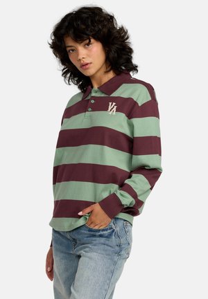 Joven con cabello rizado que lleva una camiseta de rugby a rayas verde y burdeos y jeans azul claro, posando con una mano en el bolsillo.