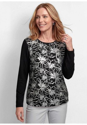 Frau mit schulterlangen blonden Haaren trägt ein schwarzes Langarmshirt mit weißem Blumenmuster und hellgraue Hose, lächelt und berührt ihr Haar.