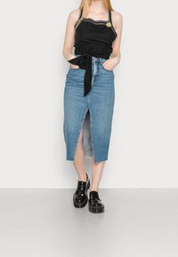 Top negro sin mangas con tirantes decorativos, cintura anudada y una falda lápiz de mezclilla azul claro con una abertura frontal. Zapatos negros con cordones.