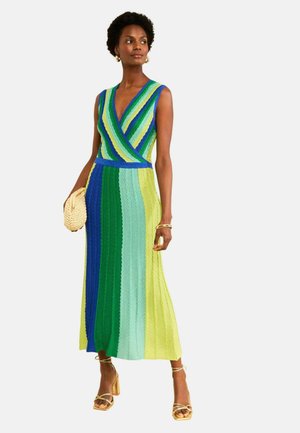 Žena nosi bezrukávové viacfarebné pruhované pletené šaty v zelených, modrých a žltých tónoch, drží pletené psaníčko a nosí zlaté sandále.