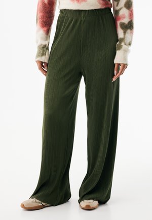Zielone spodnie z szerokimi nogawkami, z pionowymi plisami i elastycznym paskiem w talii, połączone z beżowo-brązowymi butami, prezentujące luźny krój.