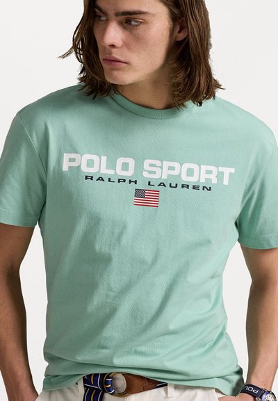 Gaišzaļš kokvilnas t-krekls ar uzrakstu "POLO SPORT RALPH LAUREN" treknām baltām burtiem un nelielu Amerikas karoga grafiku priekšpusē.