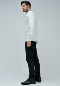 Suéter gris claro, superpuesto a una camisa azul con cuello, combinado con jeans negros y botas negras con cordones, de pie frente a un fondo gris.