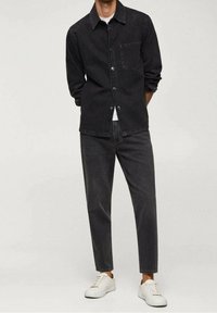 Veste chemise en denim noir avec col, poches avant et fermeture à boutons, associée à un pantalon en denim noir à coupe effilée et des baskets blanches.