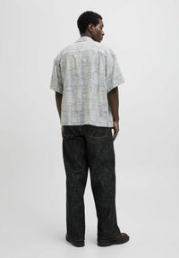Kortærmet skjorte i lysegrå med et mønstret design, parret med mørke, løstsiddende jeans og sorte sko. Bagsiden vises.