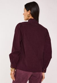 Camicia di velluto a coste bordeaux con maniche a sbuffo e scollatura arricciata. Presenta polsini con bottoni e una finitura testurizzata. Abbinata a pantaloni coordinati.