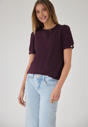 Młoda kobieta uśmiechająca się, ubrana w ciemnofioletową bluzkę z krótkim rękawem z złotymi guzikami na rękawach i jasnoniebieskie dżinsy, stojąca na jednolitym tle.