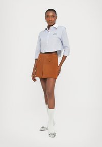 Donna con una camicia a quadri blu accorciata, una mini gonna color ruggine con bottoni, calze bianche e scarpe argentate a punta, in piedi su sfondo bianco.