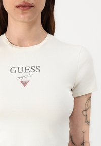 Kermanvärinen, istuva t-paita, jossa on pyöreä pääntie. Edessä on painettu "GUESS"-logo tummanharmaana sekä punainen kolmion muotoinen korostus.