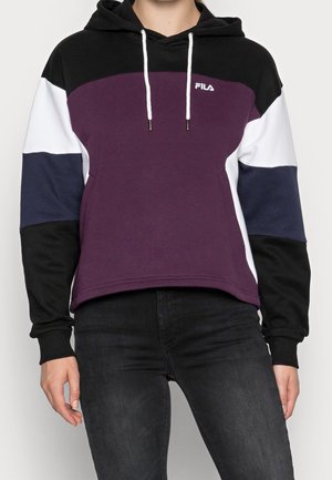 Persoon die een colourblock-hoodie draagt met zwarte, witte, marineblauwe en paarse gedeelten, met witte trekkoorden en een klein FILA-logo op de borst.