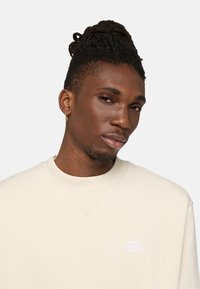 Sudadera beige de cuello redondo con ribete en el cuello y un sutil detalle de logo. Textura suave, ajuste casual y tela de peso medio.