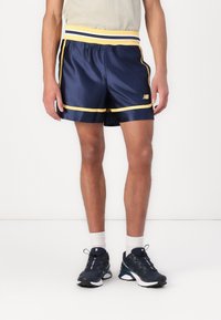Shorts de sport navy en tissu brillant avec une ceinture rayée jaune et blanche, des garnitures latérales contrastantes et un logo sur le bas devant.