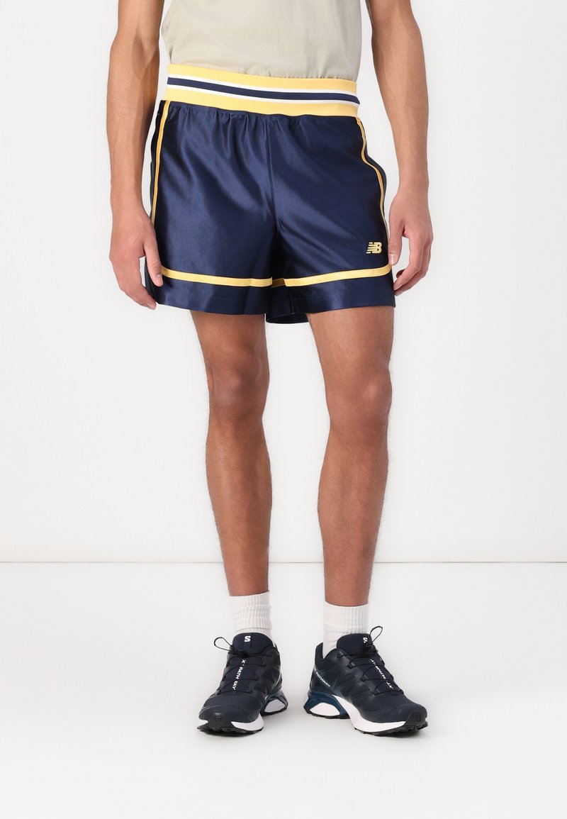 Shorts de sport navy en tissu brillant avec une ceinture rayée jaune et blanche, des garnitures latérales contrastantes et un logo sur le bas devant.