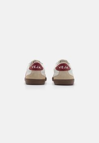 Zapatilla blanca y beige con suela de goma marrón y detalle en el talón rojo. Logo "VEJA" impreso en blanco en el talón. Materiales de ante y cuero.
