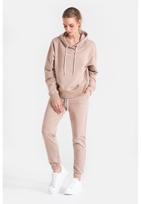 Beżowa bluza z kapturem i sznurkiem, z kieszenią kangurzą, w komplecie z dopasowanymi zwężającymi się dresami i białymi trampkami. Gładka tekstura.