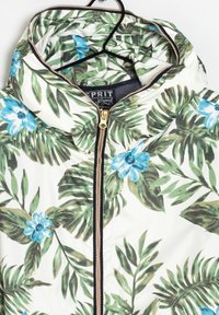 Gevoerde jas met een witte basis en een groen-blauw tropisch bloemenpatroon. Gouden ritsdetails en zwarte accenten langs de naden.