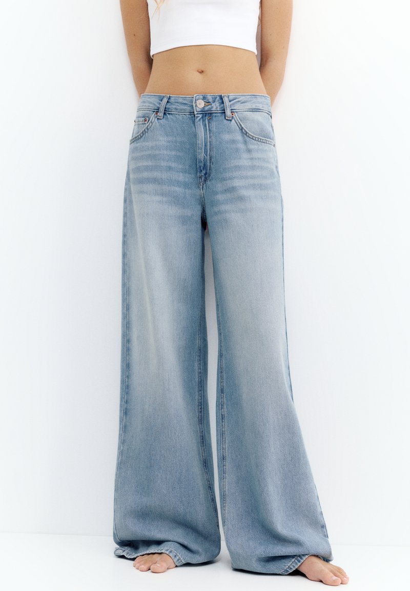 Personne portant un jean bleu clair ample à jambes larges et un haut court blanc, debout pieds nus sur un fond blanc uni.