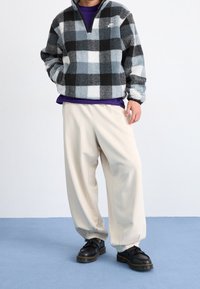 Fleece-pull med svart, grå och vit rutigt mönster, hög krage och dragkedja. Löst sittande gräddfärgade byxor och svarta skor med tjocka sulor.