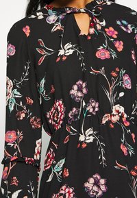 Schwarzes Blumenkleid mit langen Ärmeln, das über einen Auschnitt mit Schlüsselöffnung und eine Schleife verfügt. Der Stoff zeigt bunte Blumenmuster in verschiedenen Farbtönen.
