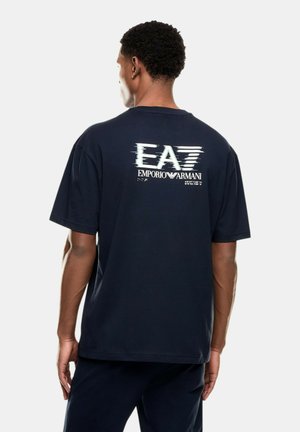 Uomo di spalle che indossa una maglietta nera con il logo e il design bianco EA7 Emporio Armani sulla parte superiore della schiena su uno sfondo uniforme.