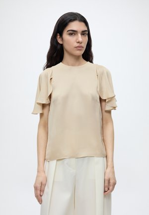 Femme aux longs cheveux foncés porte une blouse beige avec des manches courtes volantées et un pantalon blanc ample sur un fond clair uni.