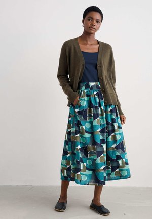 Olive green cardigan with button closure, paired with a patterned, knee-length skirt in blue, green, and navy geometric designs. Black slip-on shoes.