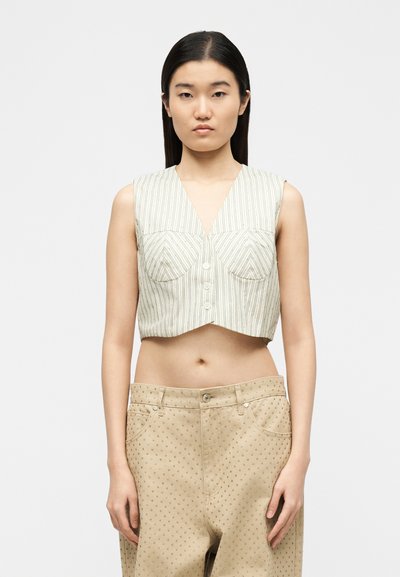 Νεαρή γυναίκα με ίσια σκούρα μαλλιά φοράει αμάνικο, ριγέ κοντό τοπ και μπεζ παντελόνι με μικρές τρουκς, πάνω σε απλό φόντο.