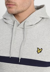 Haut de survêtement gris clair en mélange de coton, avec une bande navy sur la poitrine et un logo d'oiseau jaune sur le côté gauche. Capuche avec cordon de serrage.