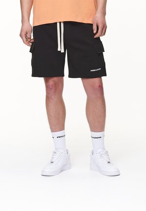 Pantalones cortos de carga negros con cordón blanco, con bolsillos laterales, combinados con zapatillas blancas y calcetines, todos mostrando el logo "Pegar".