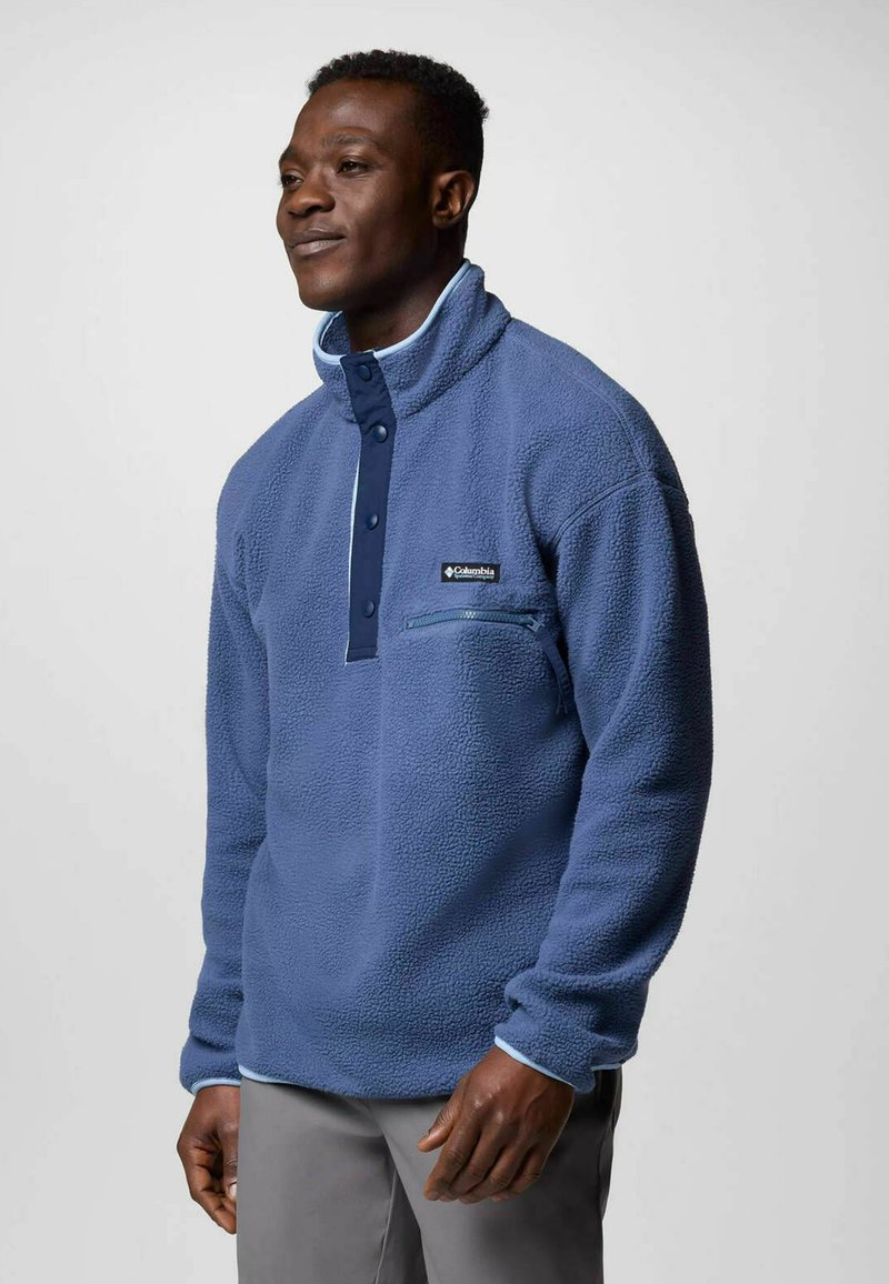 Polarowy pulower w średnim niebieskim kolorze, z zapinaną kieszenią na piersi, kołnierzem zapinanym na guziki oraz kontrastującymi ciemnoniebieskimi akcentami.