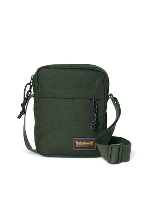 Pieni tummanvihreä olkalaukku säädettävällä hihnalla, etutaskulla vetoketjulla ja Timberlandin logolla.