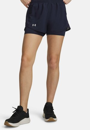 Benen met zwarte Under Armour hardloopshorts en zwarte sportschoenen met witte zolen, staand op een witte achtergrond.