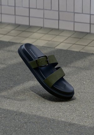 Schwarze Plateau-Pantolette mit zwei olivgrünen Riemen und einer silbernen Schnalle, ruhend auf grauem Asphalt nahe einer gepflasterten Mauer.