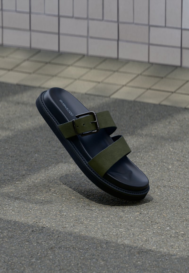 Μαύρο σαγιονάρι με δύο ελιάς πράσινες λωρίδες και ασημί αγκράφα, ακουμπισμένο σε γκρι άσφαλτο κοντά σε πλακόστρωτο τοίχο.