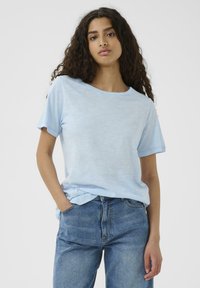 Ljusblå kortärmad t-shirt i mjukt material; avslappnad passform med rund halslinning och något upprullade ärmar. Kombinerat med blå denimjeans.
