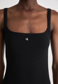Black ribbed tank top with square neckline and narrow straps, features a small white cK logo on the chest area. Smooth texture.