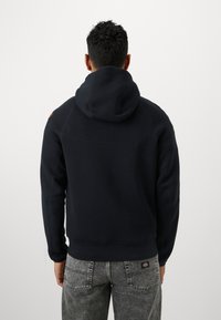 Sudadera azul marino con capucha ajustable, puños y dobladillo acanalados, que presenta un pequeño logo en el hombro izquierdo y está fabricada con una suave mezcla de algodón.