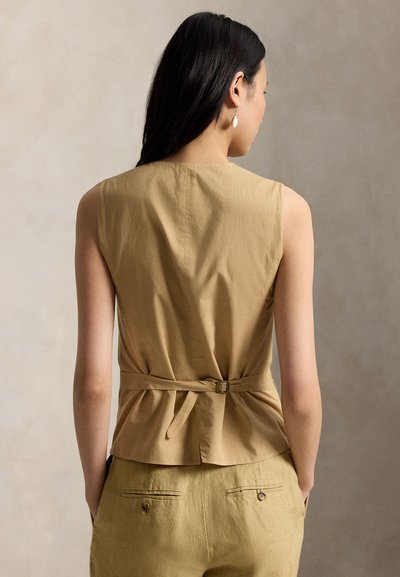 Γυναίκα με μακριά σκούρα μαλλιά που φοράει αμάνικη μπεζ μπλούζα με ρυθμιζόμενο λουρί στο πίσω μέρος και ταιριαστό μπεζ παντελόνι, στέκεται μπροστά σε ουδέτερο φόντο.