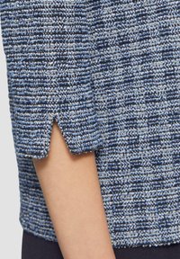Blau und weiß gemusterter Stoffoberteil mit drei Viertel-Ärmeln, das einen kleinen gewellten Ausschnitt am Bündchen aufweist.