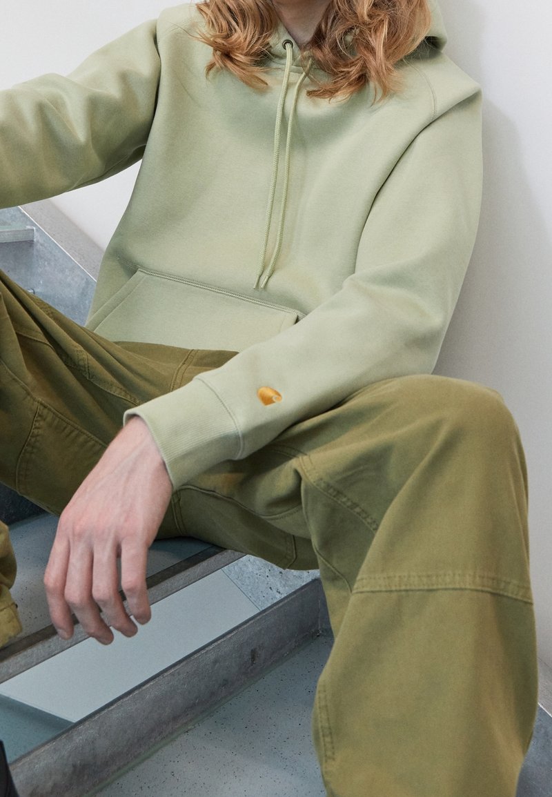 Felpa verde chiaro con tasca frontale, cappuccio con cordino e piccolo logo sulla manica, abbinata a pantaloni cargo verde oliva con dettagli cuciti.
