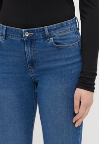 Kék farmernadrág slim fit szabással, klasszikus öt zsebes kialakítással, brass gombos záródással és diszkrét varrási részletekkel az éleken.