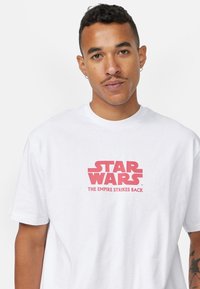 Bílý bavlněný tričko s nápisem "STAR WARS" v tučném červeném písmu, s podtitulem "IMPRÉR ZÚČTOVÁNÍ" níže v menším písmu.
