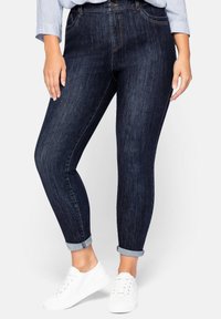 Jeans ajustados de color azul oscuro con una textura suave, que cuentan con cinco bolsillos, un cierre de botón y puños enrollados. Combinados con zapatillas blancas.