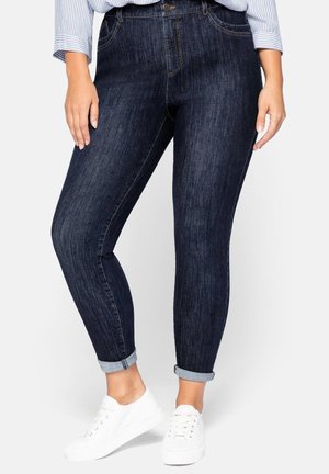 Jeans ajustados de color azul oscuro con una textura suave, que cuentan con cinco bolsillos, un cierre de botón y puños enrollados. Combinados con zapatillas blancas.