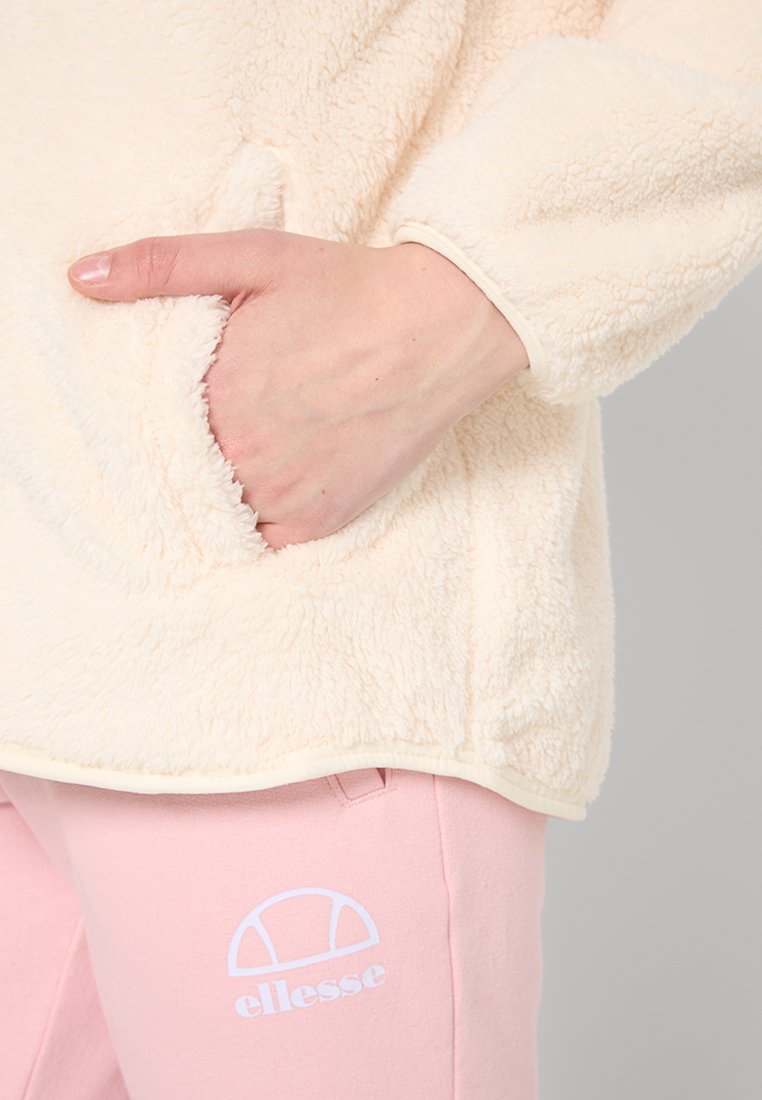 Main dans la poche d'une veste en polaire crème portée avec un pantalon de jogging rose Ellessee, montrant la texture et le logo sur le pantalon.